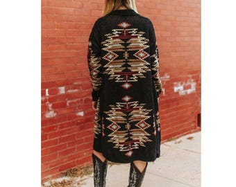 May include: A long black cardigan with a Southwestern geometric pattern in shades of brown, tan, and red. The cardigan is worn with a brown cowboy hat and black cowboy boots. The outfit is set against a red brick wall.