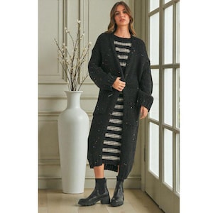 May include: A woman in a long, dark grey cardigan with colourful flecks, worn over a striped dress. The cardigan has pockets and a relaxed fit. She is also wearing black ankle boots.