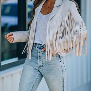 May include: A beige fringe jacket with long fringe sleeves, paired with a white top and light-wash blue jeans. The jacket has a classic blazer cut. A gold necklace is visible. The outfit is casual and stylish.