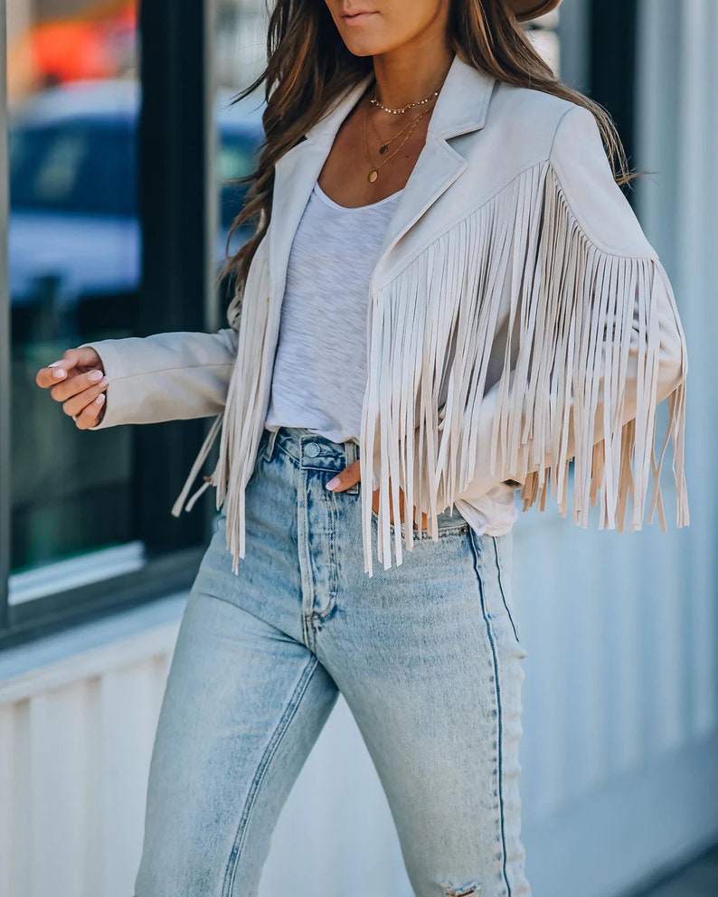 May include: A beige fringe jacket with long fringe sleeves, paired with a white top and light-wash blue jeans. The jacket has a classic blazer cut. A gold necklace is visible. The outfit is casual and stylish.