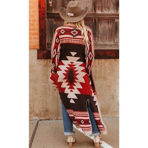 Op de afbeelding: Lange, openvallende cardigan met een geometrisch patroon uit het zuidwesten in rood, crème en bruin. De cardigan wordt gedragen met een blauwe spijkerbroek en een bruine cowboyhoed. Het ontwerp heeft ruit- en zigzagmotieven.