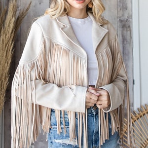 May include: A beige suede jacket with long fringe detailing on the sleeves and front. The jacket has a classic collar and is paired with denim shorts and a brown hat. The overall style is bohemian and western-inspired.