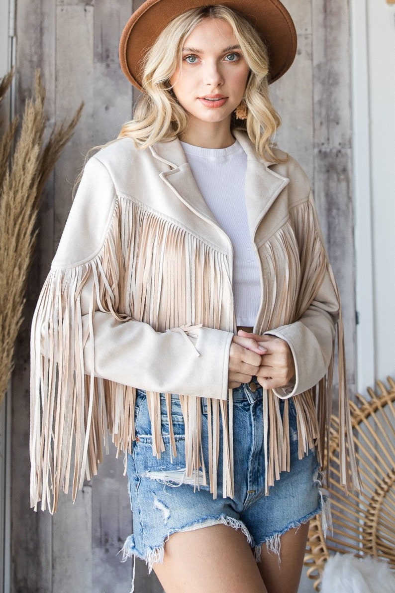 May include: A beige suede jacket with long fringe detailing on the sleeves and front. The jacket has a classic collar and is paired with denim shorts and a brown hat. The overall style is bohemian and western-inspired.