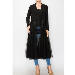 May include: A black blazer and long tulle overlay dress. The blazer has long sleeves and a tailored fit. The dress features a sequined bodice and a flowing tulle skirt. The model is wearing blue jeans and nude heels.