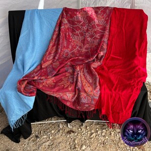 Könnte beinhalten: Drei Schals in verschiedenen Farben werden gezeigt. Ein hellblauer Schal mit Fransen, ein roter Schal und ein gemusterter roter Schal mit Fransen. Die Schals sind über einer schwarzen Oberfläche drapiert.