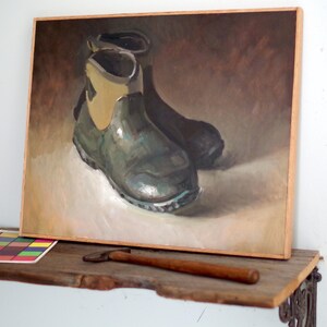 Pu&ograve; includere: Dipinto a olio di due stivali di gomma verdi con dettagli beige. L'opera &egrave; incorniciata in legno chiaro, su uno sfondo neutro. Una tabella dei colori e uno strumento di legno poggiano su una mensola di legno sottostante.