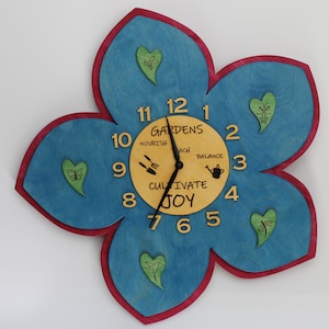 May include: A blue and yellow wooden flower-shaped clock with a red border. The clock face has the words "Gardens Cultivate Joy" and the numbers 1 through 12. There are small green hearts with images of butterflies and dragonflies around the clock face.