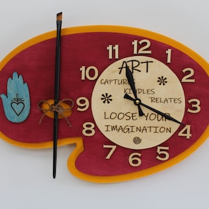 Puede incluir: Un reloj de madera con forma de paleta de artista con un esquema de color rojo y amarillo. La esfera del reloj está hecha de madera natural con los números del 1 al 12 y las palabras "ART", "CAPTURE", "KINDLES", "RELATES", "LOOSE YOUR IMAGINATION" impresas en ella. Las manecillas del reloj son negras y el reloj está configurado a las 10:50. Un pincel está unido al lado de la paleta.