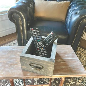 May include: A gray wooden box with a metal handle sits on a light wood table. Two black remote controls are inside the box.