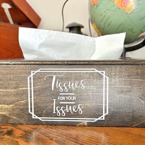 May include: A wooden tissue box with a dark stain. The box has a white graphic that reads "Tissues for your Issues". White tissues are visible at the top of the box. The box is sitting on a wooden surface.