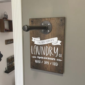 May include: A rustic wooden sign with a black metal pipe for hanging clothes. The sign reads "The Self Service Laundry Co. Open 24 Hours Wash Dry Fold".