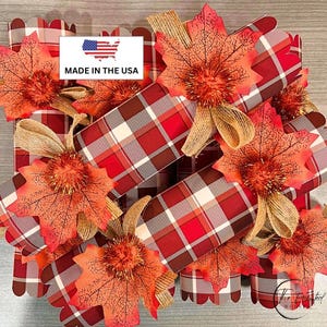 May include: A collection of cylindrical party crackers wrapped in red, white, and grey plaid paper. Each cracker is decorated with a faux orange and red maple leaf, a tan burlap ribbon bow, and a metallic orange pom-pom. A label reads MADE IN THE USA.