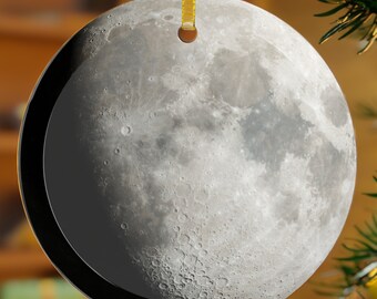 May include: A round, gray and white moon ornament with a black border. The ornament is hanging from a gold ribbon on a Christmas tree.