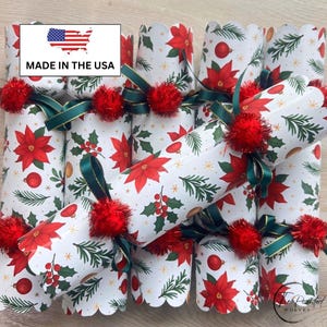 May include: A set of Christmas crackers featuring a white background with a pattern of red poinsettias, green pine branches, and red ornaments. Each cracker is tied with dark green ribbon and accented with a small, fuzzy red pom-pom.