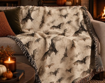 May include: A woven throw blanket featuring a repeating pattern of dark gray bats against a light beige background. The blanket has a textured appearance with fringed edges along the sides, displaying a detailed illustration of flying bats.