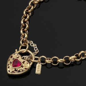 May include: Gold-toned necklace with a heart-shaped pendant. The pendant features a red gemstone and floral cutouts. The necklace has a chain-link design and a small tag that reads "18KGL".