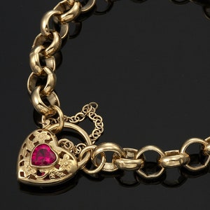 以下が含まれることがあります： 赤い宝石と花のカットアウトが特徴の、ハート型のチャームが付いたゴールドカラーのブレスレット。 ブレスレットは、ロブスタークラスプ留めの太いチェーンデザインです。 ジュエリーは黒い背景に表示されています。