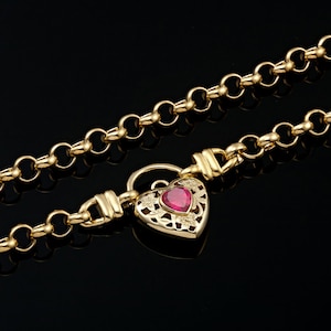 May include: Gold-coloured necklace with a heart-shaped pendant. The pendant has a red gemstone centre and detailed floral cutouts. The chain is made of interlocking circles, creating a classic and elegant look. The necklace is displayed against a black background.