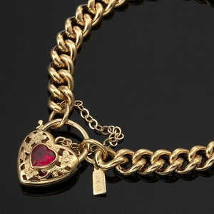 May include: A gold necklace with a heart-shaped pendant. The pendant has a red gemstone and floral cutouts. The necklace features a thick chain design and a small tag that reads "18K GL".