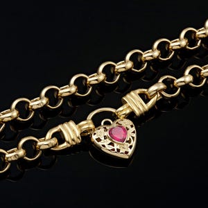 以下が含まれることがあります： 金色のチェーンネックレス、ハート型のペンダント付き。ペンダントの中央には赤い宝石があり、花の透かし模様で飾られています。ネックレスは黒い背景に表示されています。