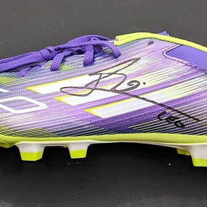 May include: A purple and lime green soccer cleat featuring a gradient striped pattern, white laces, and the text F50 printed on the side. The exterior of the shoe displays a black ink signature across the midsection, with lime green studs.