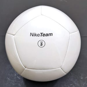 以下が含まれることがあります： 黒い文字でNikeTeamと書かれ、SIZE 5という文字が入った円形のロゴがプリントされた、白いサッカーボール。縫い合わされたパネルで構成されており、滑らかで光沢のある仕上げの球体で、標準的なスポーツ用途向けに設計されています。