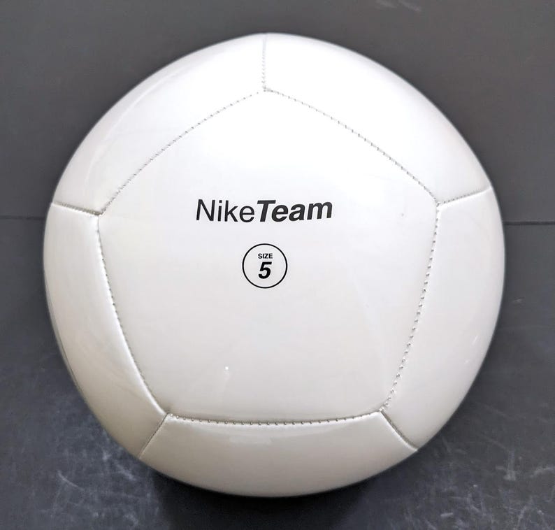 以下が含まれることがあります： 黒い文字でNikeTeamと書かれ、SIZE 5という文字が入った円形のロゴがプリントされた、白いサッカーボール。縫い合わされたパネルで構成されており、滑らかで光沢のある仕上げの球体で、標準的なスポーツ用途向けに設計されています。