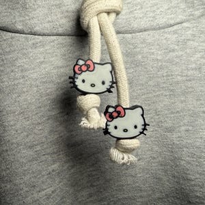May include: A heather gray hooded sweatshirt featuring two thick, cream-colored cotton drawstrings. Each drawstring is adorned with a white Hello Kitty character charm, complete with a pink bow, black whiskers, and black eyes, secured by knots.