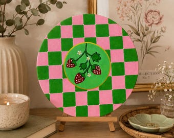 May include: A circular wooden plaque featuring a vibrant pink and green checkerboard pattern. In the center, a circular green medallion displays a hand-painted illustration of three red strawberries with white seeds, green leaves, and small white flowers.