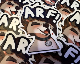May include: A pile of stickers depicting a cartoon wolf with brown fur, wearing a black collar with a tag that says "Bitch". The wolf is smiling with its mouth open, and the word "ARF!" is written above its head in white letters.