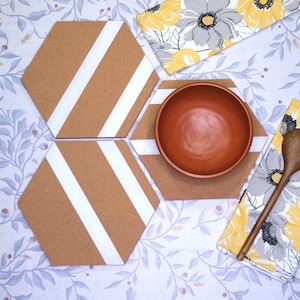 May include: Four hexagonal cork trivets with white stripes, a terracotta bowl, and a wooden spoon on a white tablecloth with a floral pattern.