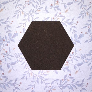 Puede incluir: Un tablero de corcho hexagonal de color marrón claro y superficie texturizada. El tablero de corcho está colocado sobre un fondo blanco con un delicado patrón floral.