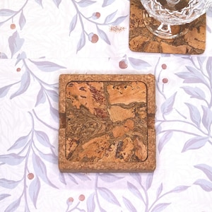 May include: Square cork coasters with a natural wood grain pattern. The coasters have a raised border and are designed to protect surfaces from water rings. The coasters are placed on a white tablecloth with a floral pattern.