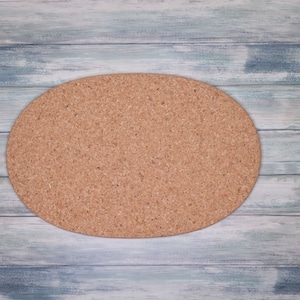 Può includere: Un sottopentola ovale in sughero, di colore marrone naturale, poggia su una superficie di legno bianco e azzurro chiaro. Questo sottopentola è progettato per proteggere le superfici dal calore ed è un semplice accessorio da cucina.