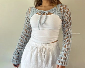 May include: A light blue crochet shrug with a tie at the neck. The shrug is worn over a white tank top and white pants.