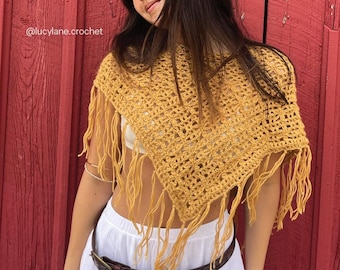 May include: Mustard yellow crochet poncho with fringe detail. The open weave design is visible. The background is a red wooden wall. The text "@lucylane.crochet" is visible.