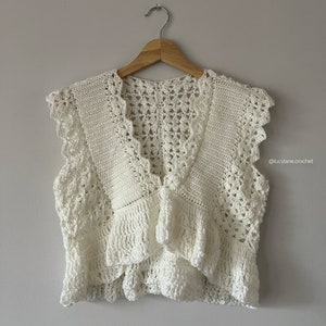 Puede incluir: Un chaleco blanco de ganchillo con un escote en V profundo y ribete de volantes. El chaleco tiene un delicado patrón de encaje y está colgado en una percha de madera. @lucylane.crochet