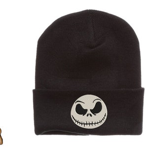 May include: Black beanie featuring an embroidered Jack Skellington face. The beanie has a folded cuff and a round patch with the character's face. The text "BOO BEAR DESIGNZ whimsical embroidery designs" is on the left side.