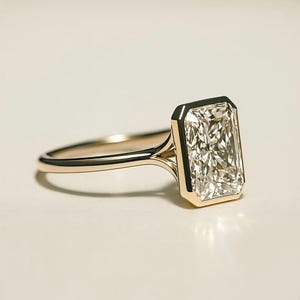 May include: A gold ring featuring a large, rectangular-cut diamond. The diamond is set in a bezel setting, and the band is a simple, polished gold. The ring is displayed on a white surface.