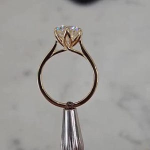 Puede incluir: Un anillo de oro con una piedra preciosa redonda de talla brillante sujeta por un engaste de seis garras que recuerda a una flor de tulipán. El aro es fino y pulido, con un tono oro amarillo cálido y una piedra transparente facetada.