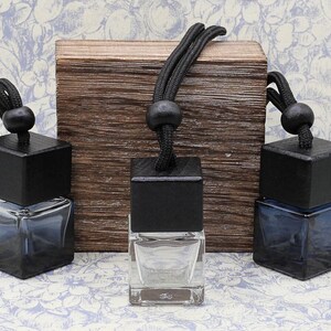 May include: Three square glass bottles with black wooden caps and black string hangers. The bottles are filled with a clear liquid and are displayed on a wooden surface.