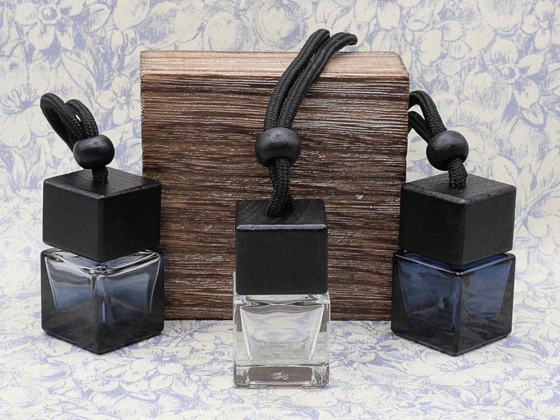 May include: Three square glass bottles with black wooden caps and black string hangers. The bottles are filled with a clear liquid and are displayed on a wooden surface.