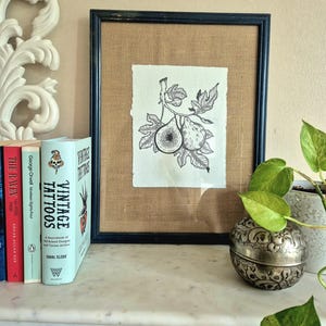 May include: Black framed fig art print. The black and white illustration depicts figs on a branch with leaves. The artwork is matted with a burlap-like material. The print is displayed on a white marble mantel with books and a decorative silver container.