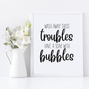 以下が含まれることがあります： 白い額縁に入ったプリントで、黒い文字で「Wash away those troubles have a soak with bubbles」と書かれています。プリントは白い表面に置かれており、白いピッチャーと白い花が飾られています。