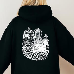 May include: Black hoodie with a white graphic on the back. The graphic features an illustration of a deity riding a horse, with the text "KRISHNA IS MY COPILOT" in a stylized font. The design includes a wheel and floral elements.