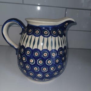 May include: A ceramic pitcher featuring a cream-colored base with a hand-painted pattern of dark blue peacock eye motifs and vertical teal stripes. The vessel has a curved handle, a wide spout, and a rounded body with a glossy finish.