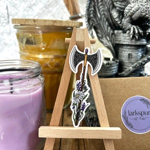 May include: A sticker featuring a black and silver axe with a purple gem on the handle. The axe is surrounded by white and purple flowers. The sticker is on a small wooden easel. The text "larks pur art studio" is on a brown paper tag.