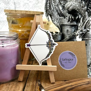 May include: A white sticker with a purple and green floral design of a bow and arrow. The sticker is on a small wooden easel. The sticker has the text "larksour art studio" in a circle.
