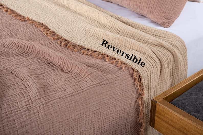 Puede incluir: Una manta reversible con tejido de gofre texturizado y ribete de flecos. Un lado es de color beige claro y el otro lado es de color marr&oacute;n claro. La manta se muestra doblada en una cama.