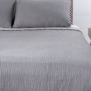 May include: Gray textured waffle weave quilt with a slightly frayed edge. The quilt is folded back on a bed with two gray pillows.